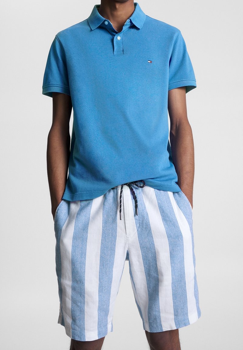 Homme portant un polo bleu à manches courtes et un short blanc avec des rayures verticales bleu clair, les mains dans les poches, se tenant devant un fond uni.