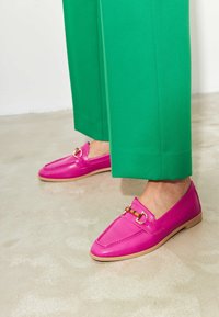 Roze leren loafers met een gouden gespaccent. Ze hebben een klassiek ontwerp en een platte tan zool, gecombineerd met groene broek.