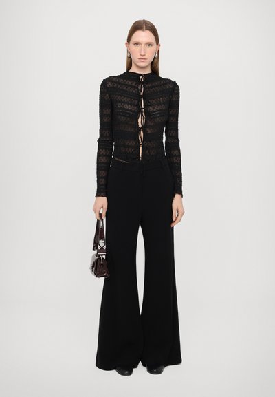 Zwart aangesloten kanten top met lange mouwen en een voorzijde met vetersluiting, gecombineerd met zwarte wide-leg broek. Model houdt een kleine bordeauxrode handtas vast.