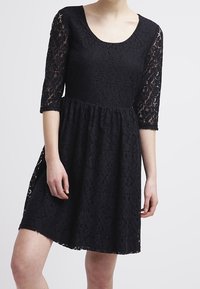 Schwarzes Spitzenkleid mit Dreiviertelärmeln und runder Ausschnitt. Verfügt über einen ausgestellten Rock und ein filigranes Spitzenmuster durchgehend.