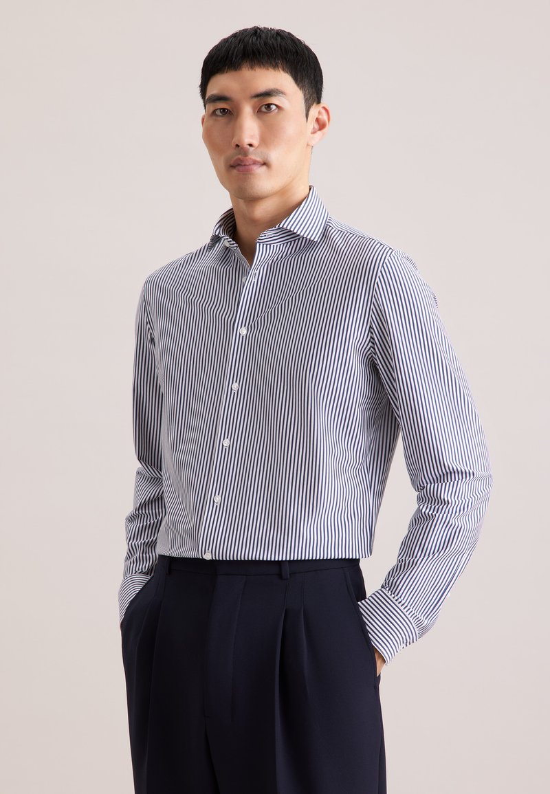 Homme portant une chemise boutonnée à rayures verticales bleues et blanches, rentrée dans un pantalon noir plissé, mains dans les poches, fond neutre.