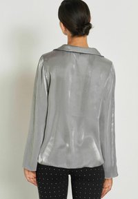 Woman with dark hair in a low bun wears a shiny silver long-sleeve blouse and black pants with small white polka dots, seen from the back.