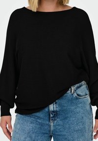 Modell som bär en löst sittande svart ribbat topp och blå högmidjade jeans, som visar en avslappnad, casual stil.