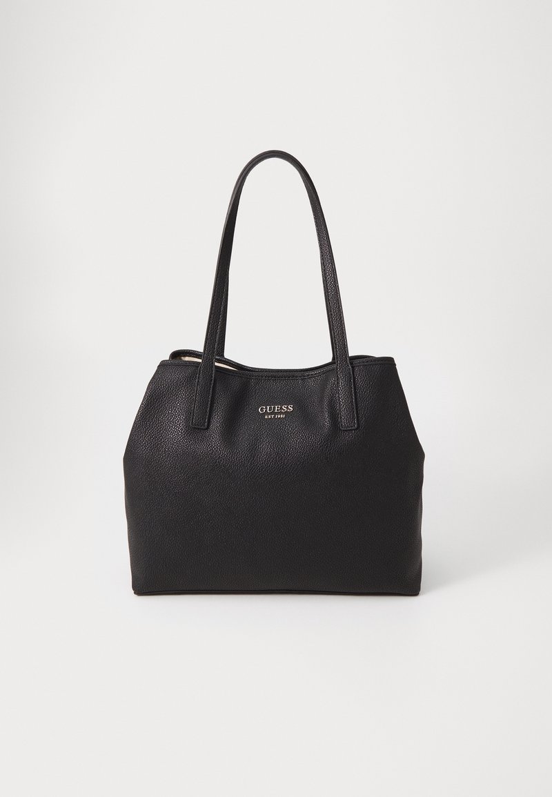 Sac cabas Guess en cuir noir avec deux poignées et petit logo doré à l'avant, debout sur fond blanc.