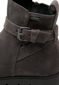 Bottines en suede grises avec une surface texturée, dotées d'une sangle décorative avec une boucle métallique et d'une semelle noire épaisse.