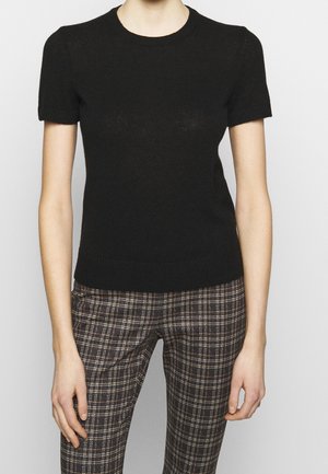 Personne portant un haut en maille noir à manches courtes et un pantalon à carreaux taille haute sur un fond clair uni.