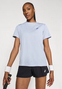 Απαλή μπλε αθλητική μπλούζα με κοντά μανίκια, στρογγυλή λαιμόκοψη και μαύρο λογότυπο. Συνδυασμένη με σκούρες βερμούδες και λευκή βραχιόλι.