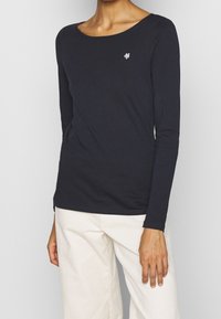 Nainen, jolla on päällä musta pitkähihaisa paita, jossa on pieni valkoinen logo rinnassa, ja vaaleanbeige housut, taustalla yksinkertainen tausta.