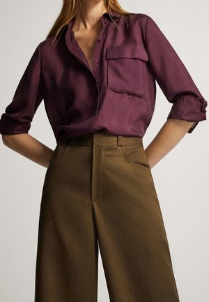 Femme portant une chemise violette à manches retroussées, rentrée dans un pantalon marron taille haute et jambes larges, debout les mains sur les hanches.