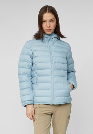 Ung kvinde iført en lyseblå dunjakke og beige bukser, stående med den ene hånd i lommen mod en ensfarvet grå baggrund.