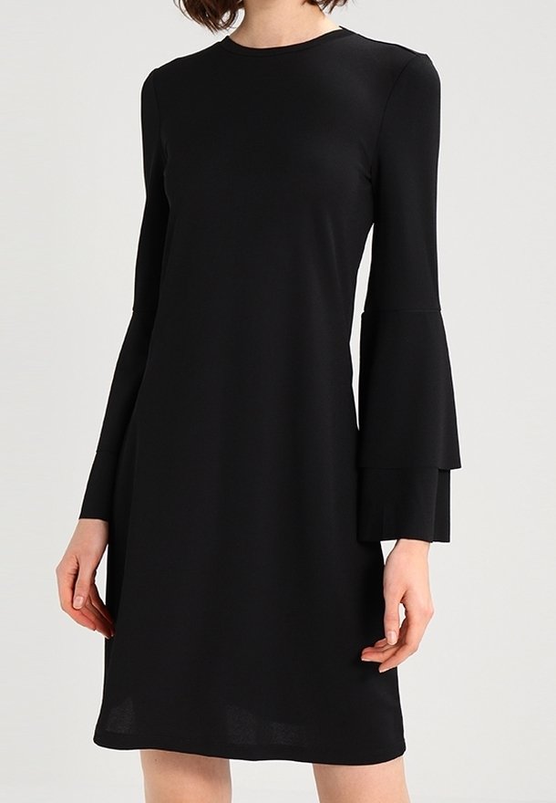 Femme portant une robe noire à longueur genou avec des manches longues évasées à étages et un col rond sur un fond uni.