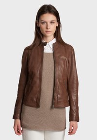 Bruine leren jack met een rits aan de voorkant, slim fit en een ronde kraag. Gelaagd over een beige trui. Soepele textuur en op maat gemaakt ontwerp.