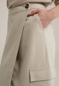 Beige skräddarsydda byxor, tillverkade av mjukt tyg. Har en framlomma och en dekorativ knapp, med en strukturerad design och rena linjer.