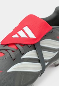Gray soccer shoe with red and white accents, featuring a textured upper, flexible strap, and contrasting detailing on the side.