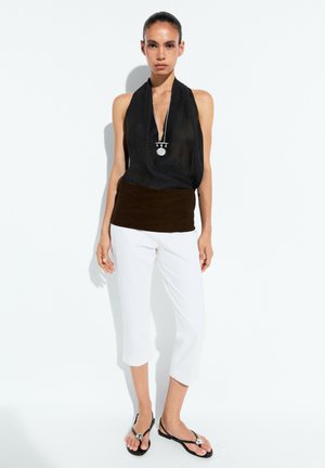 Femme portant un haut noir sans manches, un pantalon blanc court, des sandales noires et un collier pendentif en argent, debout devant un fond blanc.