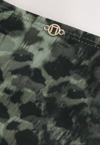 Gros plan sur un tissu tie-dye vert foncé et noir avec un petit emblème métallique rond argenté cousu près du bord.