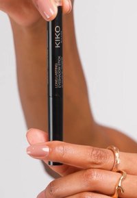 Musta sylinterimäinen luomivärikynä, jossa lukee "KIKO Long Lasting Eyeshadow Stick", käsissä vaaleanpunaiset kynnet ja kullanväriset sormukset.