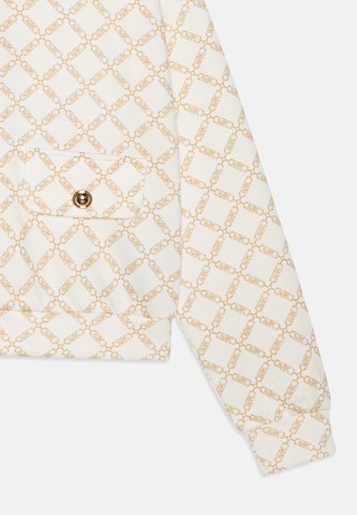 Valkoinen collegepaita, jossa on monimutkainen beige timantinmuotoinen kuvio, etutasku ja kultaiset napit hihansuissa ja edessä.