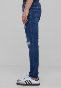 Temno modri jeans s ožjim krojem, ki imajo obrabljene detajle na kolenih, obuti z belimi in sivimi športnimi čevlji.