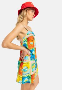 Colorful dress with a postcard print featuring tropical themes. Thin straps, A-line shape, lightweight fabric. Red bucket hat as an accessory.