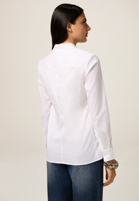 Camicia bianca a maniche lunghe con colletto, caratterizzata da una texture liscia e un taglio dritto. Abbinata a jeans blu a gamba larga e a un braccialetto a fantasia.