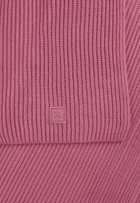 Écharpe tricotée en rose pâle, avec une texture côtelée et une petite étiquette carrée avec un motif floral dans un coin.