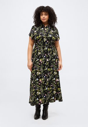 Frau mit lockigem Haar, die ein langes schwarzes Blumenkleid mit kurzen Ärmeln und schwarzen Stiefeletten trägt und vor einem einfachen Hintergrund steht.