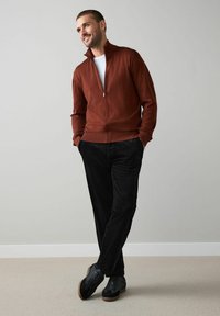 Barna cipzáros pulóver, fehér póló alatta, fekete kordnadrág, és fekete tornacipő barna talppal. Sima textúra, modern szabás.