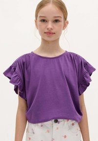 Lilla kortærmet T-shirt med flæser ved skuldrene, afslappet pasform og blødt bomuldsstof. Sammen med trykte lyse bukser.