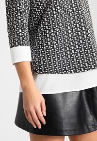 Svartvitt mönstrad topp med vit manschett och fåll, tillsammans med en svart läderkjol. Texturerad yta, stilren design.