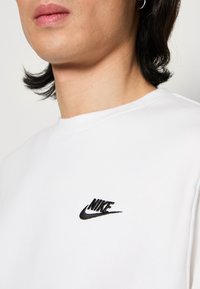 Bílá mikina vyrobená z bavlny, s černým vyšitým logem Nike na levé části hrudi. Kulatý výstřih, mírně viditelná textura.
