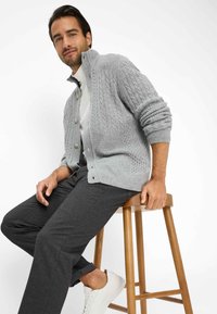 Grijze kabelgebreide cardigan met hoge kraag en knoopsluiting, gedragen over een wit shirt, gecombineerd met donkere broeken en witte sneakers.