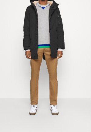 Sort parka med pelsforet hætte, lagdelt over en grå sweater med blå og grønne striber, kombineret med kaki bukser og hvide sneakers.