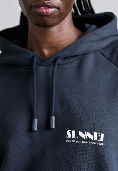 Sweatshirt marine en mélange de coton. Il présente un logo sur le devant avec "SUNNEI" et un slogan. Les cordons au niveau du col sont gris avec des embouts en plastique.