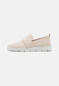 Zapato de ante beige tipo slip-on con una amplia correa de tela en la parte superior, con suela de goma blanca y textura suave.