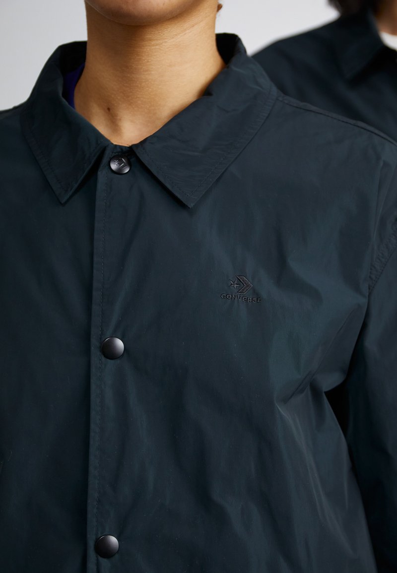 Giacca nera in nylon con chiusura a bottoni e colletto a punta. Presenta un piccolo logo in rilievo sul petto e una texture liscia e opaca.