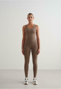 Brązowy sportowy top na ramiączkach i legginsy z wysokim stanem, oba wykonane z elastycznego materiału. Białe buty sportowe z czarnymi akcentami dopełniają całość.