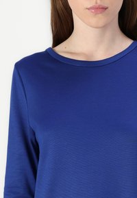 Pitkähihaisessa sinisessä paidassa on sileä kangas. Siinä on pyöreä pääntie ja istuva siluetti, jossa on hienovarainen tekstuuri.