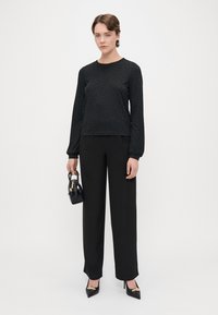 Haut noir à manches longues avec un motif texturé, associé à un pantalon noir taille haute et des chaussures à talons pointus ; tenant un petit sac à main noir.