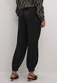 Pantaloni neri a sigaretta, larghi e con polsini elasticizzati, una sottile texture e un dettaglio con tasca sul retro. Indossati con scarpe col tacco nere.