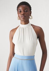 Top à col halter en tissu crème avec un design froncé et sans manches, associé à une jupe plissée bleu clair et une ceinture rehaussée d'or.