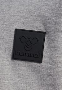 Sort gummipatch med stiliseret bi-logo og "hummel" tekst på grå strikket stof baggrund.