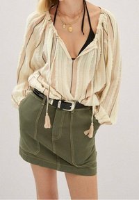 Blouse rayée beige clair avec des liens à glands, associée à une jupe en denim verte olive avec des poches avant et des coutures contrastées.