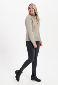 Beige vadderad jacka med hög krage, dragkedja och långa ärmar, i kombination med mörka jeans och svarta ankelstövlar.