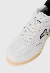 Zapatilla deportiva blanca con parte superior de cuero texturizado, perforaciones para ventilación y suela de goma. Presenta detalles y cordones en negro.