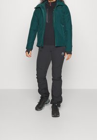 Teal vattentät jacka med huva över en svart långärmad tröja, tillsammans med svarta byxor. Har justerbara ärmslut och logodetaljer.