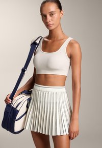 Ljus beige ribbad crop top som matchas med en plisserad ljus kjol med marinblå detaljer. Modellen håller en marinblårandig duffelväska.
