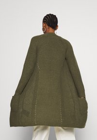 Pitkä, ylisuuri oliivinvihreä neuletakki, joka on valmistettu tekstuurilla varustetusta neulekudoksesta. Takissa on ribbireunukset ja sivutaskut.