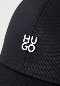 Black cap made of textured fabric featuring a silver logo pin that reads "HUGO." Shows detailed stitching on the brim and crown.
