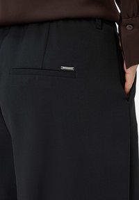 Pantalons noirs ajustés avec une texture lisse, dotés d'une poche arrière et d'une étiquette en métal argenté sur la poche pour le branding.